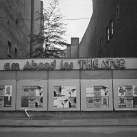 Schwarz-Weiß-Fotografie einer Plakatwand über der eine Leuchtreklame mit dem Schriftzug "am Abend ins Theater" hängt